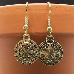 Puede incluir: Pendientes de brújula dorados con un diseño de estrella y las letras N, S, E y W alrededor del borde.