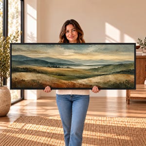 May include: A panoramic landscape painting in a black frame featuring rolling hills, distant mountains, and a soft, hazy sky in muted tones of green, gold, and brown. The artwork depicts a wide, serene valley with layered terrain and light.