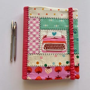 May include: A patchwork journal with a pink typewriter design and a blue heart. The cover incorporates gingham, heart, and strawberry patterns. Two silver pens are positioned beside the journal. The journal has a pink, ruffled spine.