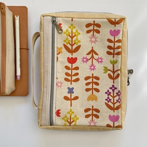 以下が含まれることがあります： 赤、黄、ピンク、紫の花が咲くカラフルな花柄のベージュ色のジッパーケース。お揃いのペンケースと茶色の革製ノートが左側にあります。