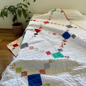 May include: A white quilt with a patchwork design featuring squares of various colours and patterns, including red, blue, green, black, and white. The quilt is folded and draped over a bed.