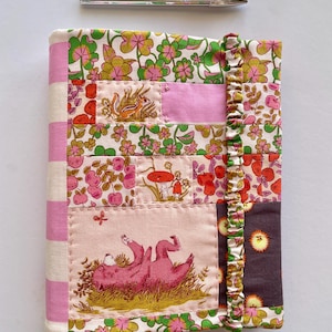May include: A patchwork fabric journal with a floral and whimsical design in pink, green, and orange. The spine is pink and white striped, and a ruffled edge adds detail. Two silver pens are above the journal. The journal is a unique, handcrafted item.