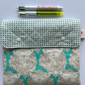 以下が含まれることがあります： 白と緑のチェック柄のフラップが付いたターコイズブルーの布製ポーチ。ポーチには、ターコイズブルーの背景に白いライオンの顔が繰り返し描かれています。ポーチのフラップには白いボタンが2つ付いています。ポーチの上に、マルチカラーのペンが2本置かれています。