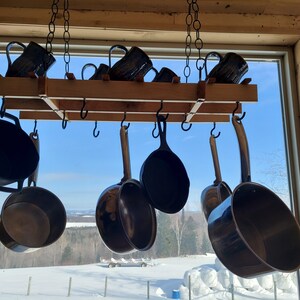 May include: A wooden pot rack with black metal hooks hanging from the ceiling. The rack is filled with various sized black pots and pans. The rack is in front of a window with a view of a snowy landscape.