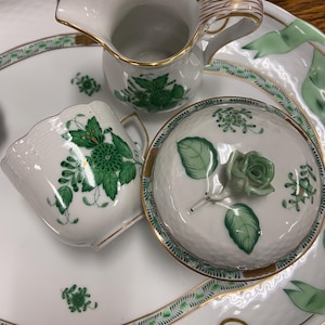Puede incluir: Un juego de té de porcelana blanca con diseños florales verdes. El juego incluye una jarra, un azucarero con tapa y una taza. El juego está en una bandeja de porcelana blanca con un borde verde y dorado.