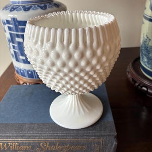 May include: A white hobnail glass bowl with a pedestal base. The bowl has a textured surface and a ruffled rim. It sits atop a blue book with gold lettering. In the background are blue and white decorative jars.