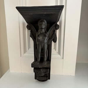 May include: A dark-colored wall bracket featuring a central sphinx figure with wings and a flat top shelf. The decorative support element displays a sculpted human-like head and feline body, finished in a matte black tone with visible wear.