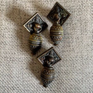 Może przedstawiać: Trzy antyczne mosiężne gałki do szafek z ozdobnymi wzorami. Każda gałka ma okrągłą, fakturowaną podstawę z ozdobnym wierzchołkiem.