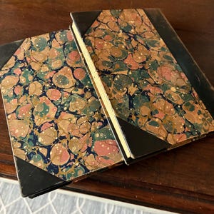 May include: Two antique books with marbled covers in pink, green, and gold. The corners and spines are black. The books are slightly open, revealing the aged, yellowed pages.