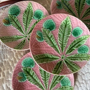 May include: Three round ceramic plates featuring a raised, embossed design of green chestnut leaves and spiky green burrs set against a textured, pale pink background. Each plate displays a consistent botanical pattern with detailed leaf veins.