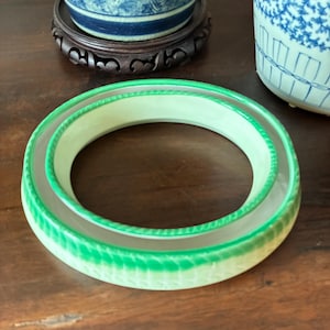 May include: A light green and white ceramic bracelet with a textured, ridged design. The bracelet is circular and appears to be hollow. The bracelet is sitting on a wooden surface. Decorative items are in the background.