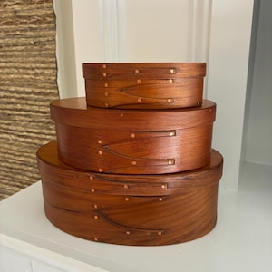 May include: A set of three oval-shaped wooden boxes stacked vertically. Each box features a smooth, medium-brown wood grain finish and is secured with small, evenly spaced copper-coloured metal tacks along the overlapping side seams.