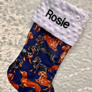 Puede incluir: Una media de Navidad azul con un puño de felpa blanca que lleva el nombre "Rosie" bordado en negro. La media está decorada con un patrón de perros dachshund negros y marrones.
