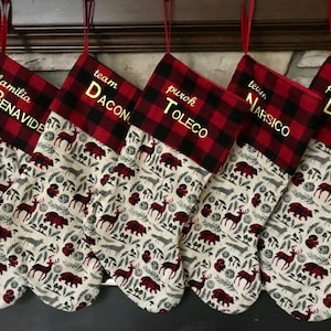 May include: Five red and black buffalo plaid Christmas stockings with a woodland pattern. The stockings are personalized with the names "familia Benavide", "team Dacon", "purok Toledo", "team Narsico", and "familia Duroy".
