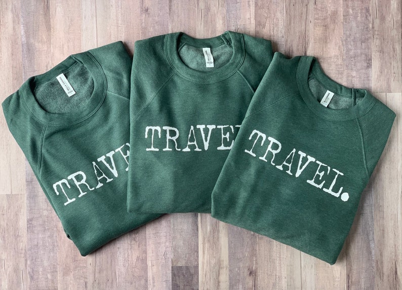 May include: Three green sweatshirts with the word "TRAVEL" printed in white on the front. The sweatshirts are folded and stacked on top of each other.