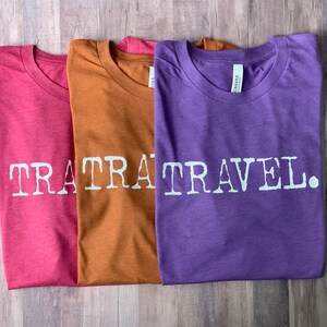 May include: Three t-shirts with the text "TRA TRAVEL." printed on them. The shirts are folded and stacked on top of each other. The top shirt is purple, the middle shirt is brown, and the bottom shirt is pink.