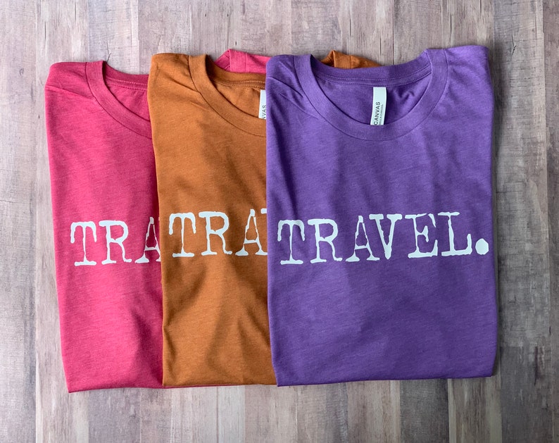 May include: Three t-shirts with the text "TRA TRAVEL." printed on them. The shirts are folded and stacked on top of each other. The top shirt is purple, the middle shirt is brown, and the bottom shirt is pink.