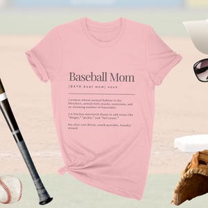 以下が含まれることがあります： 「Baseball Mom」の文字と定義が書かれた薄いピンク色のTシャツ。野球のバット、野球ボール、グローブ、サングラス、野球帽も画像に写っています。