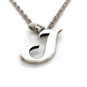 May include: A silver-toned necklace featuring a pendant in the shape of the letter "J". The pendant is a capital letter with a rounded serif design. The chain is a delicate, twisted rope style, and the background is white.