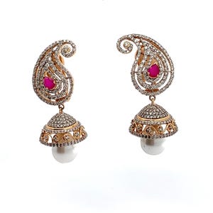Può includere: Un paio di orecchini jhumka dorati con una parte superiore a forma di paisley incastonata con pietre chiare e una pietra rosa centrale a goccia. Una base a campana coperta di pietre chiare pende sotto, rifinita con una singola perla bianca.