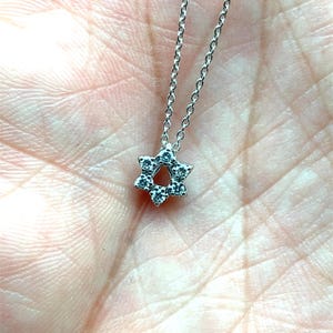 May include: A delicate silver necklace featuring a Star of David pendant. The pendant is encrusted with small, sparkling gemstones. The necklace is displayed against a soft, neutral background.