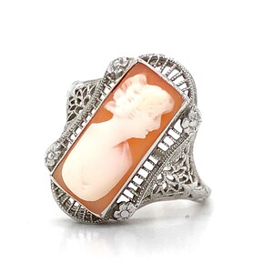 May include: A rectangular cameo ring featuring a carved profile of a person in light cream against an orange background. The setting is made of silver-toned metal with intricate filigree detailing and small floral accents at the corners.