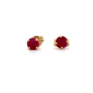 May include: A pair of stud earrings featuring round, vibrant red gemstones set in gold. The earrings have a classic design, with the gemstones held in place by four prongs. The gold setting adds a touch of elegance to the jewelry.