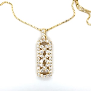 Puede incluir: Un collar dorado con un colgante rectangular. El colgante está adornado con numerosas piedras preciosas pequeñas y claras dispuestas en un patrón floral. La cadena es una fina cadena de caja dorada.