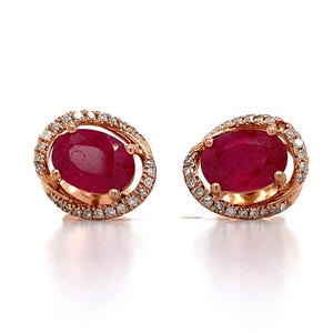 May include: A pair of oval-shaped ruby earrings with a halo of small diamonds set in rose gold. The rubies are a deep red color, and the diamonds sparkle around them. The earrings are displayed on a white background.