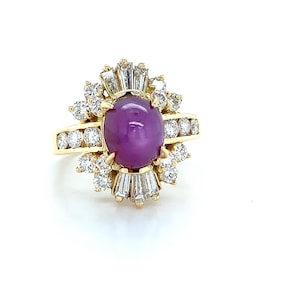 Op de afbeelding: Een gouden ring met een grote, ovale, paarsrode edelsteen in het midden, omringd door kleinere, heldere edelstenen. Het ontwerp van de ring omvat een halo van ronde en baguette-geslepen stenen, wat een sprankelende, elegante uitstraling creëert. De ring is gezet op een gouden band.