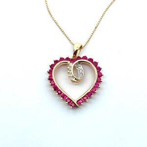 Puede incluir: Un collar de oro con un colgante en forma de corazón. El colgante está delineado con piedras preciosas rojas y presenta una sección curva más pequeña con piedras preciosas blancas. El collar está sobre un fondo blanco.