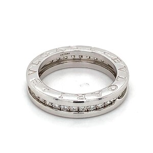 May include: A silver ring with the word "BVLGARI" engraved around the outer band. The ring features a row of small, clear gemstones set within a channel. The ring is on a white background.