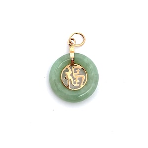 Könnte beinhalten: Ein runder, hellgrüner Jade-Anhänger mit einem goldfarbenen Rahmen und Bügel. Der Rahmen hat ein ausgeschnittenes Design eines chinesischen Schriftzeichens. Der Anhänger hängt an einer goldfarbenen Schlaufe.