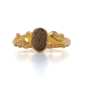 May include: A gold ring featuring an oval-shaped center plaque engraved with an intricate, stylized monogram of the letters E and L. The band displays ornate, raised scrollwork detailing on both sides of the central oval element.