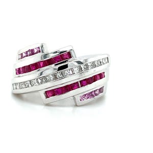 May include: A silver ring with a geometric design. The ring features alternating rows of square-cut rubies and clear, square-cut gemstones. The gemstones are set in a channel setting, creating a bold and modern look. The ring is set against a white background.