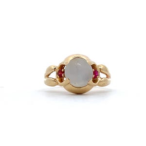 May include: A gold ring featuring a large, oval, light-colored stone in the center, flanked by two smaller red stones. The ring has a unique, ornate design with curved accents on the sides, set against a white background.