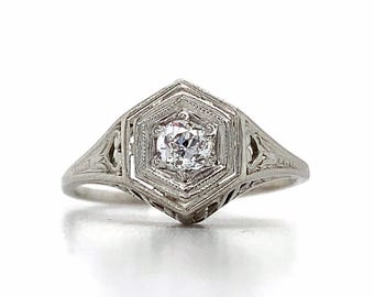 May include: A vintage white gold ring with a hexagonal setting. The ring features a central, round-cut diamond and intricate filigree detailing on the band and setting. The ring is set against a white background.