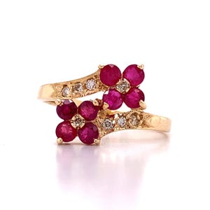Peut inclure: Une bague en or ornée de deux grappes en forme de fleur composées de rubis ronds rouge foncé. Chaque grappe de rubis est accentuée par de petits diamants ronds transparents sertis le long de l'anneau en or, qui s'incurve pour relier les deux motifs.