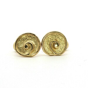 May include: Two gold-colored, spiral-shaped earrings. Each earring features a textured surface and a raised spiral design in the center. The earrings are set against a white background, highlighting their metallic sheen.