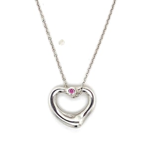 Può includere: Una collana a catena in metallo color argento con un ciondolo a forma di cuore aperto. Il ciondolo è impreziosito da una singola piccola pietra preziosa rotonda di colore rosa vivace, incastonata al centro superiore della cornice a cuore.