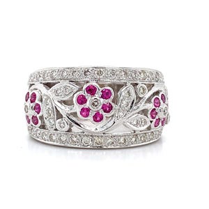 May include: A white gold ring with a floral design. The ring features a band of small diamonds and a central design of flowers with pink gemstones. The flowers are surrounded by leaves and additional diamonds.