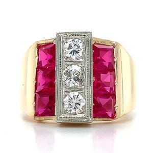 May include: A gold ring featuring a central vertical white metal bar set with three round clear stones. Flanking the central bar are two vertical rows of three square-cut vibrant red stones each, set into a polished gold band with a smooth finish.