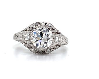 Può includere: Un anello di fidanzamento in argento con una grande pietra centrale di diamante rotonda e trasparente. L'anello presenta un'intricata filigrana e diamanti più piccoli sulla fascia. L'anello è posto su uno sfondo bianco.