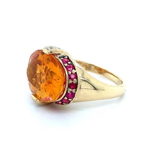 May include: A gold ring with a large, oval-cut orange gemstone. The ring is embellished with small, round red gemstones and a small cluster of diamonds. The band is a polished gold colour.