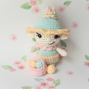 Könnte beinhalten: Eine gehäkelte Puppe, die ein pastellfarbenes blaues und gelbes Kleid und einen Hut mit einer Krone trägt. Die Puppe hält einen rosa Korb mit gelben und blauen Eiern.