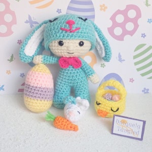 May include: A crochet doll wearing a turquoise bunny hat and suit with a pink bow tie, holding a pastel striped egg. Beside it are a small white crochet bunny head with an orange carrot, a yellow chick basket, and a sign reading Uniquely Tangled.