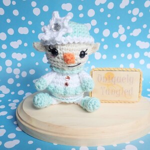 May include: A crocheted snowman figurine wearing a light blue and white hat with a sparkly snowflake on top. The figurine is sitting on a wooden base with a sign that reads "Uniquely Tangled".
