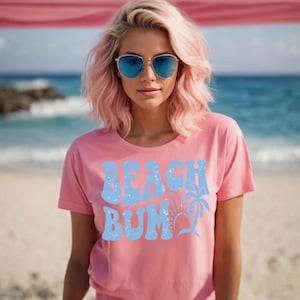 May include: A pink t-shirt with the words "BEACH BUM" in light blue, along with a palm tree and sun graphic. The person in the image is wearing blue sunglasses and has pink hair. The background is a beach scene.