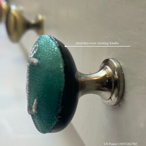 May include: A teal blue and black knob with a white embroidered design. The knob is designed to stretch over existing knobs.