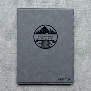 May include: Gray leatherette journal with a black logo of a mountain range and river with the text "Sage River Development". The name "Jeff P. Turk" is embossed in black at the bottom.